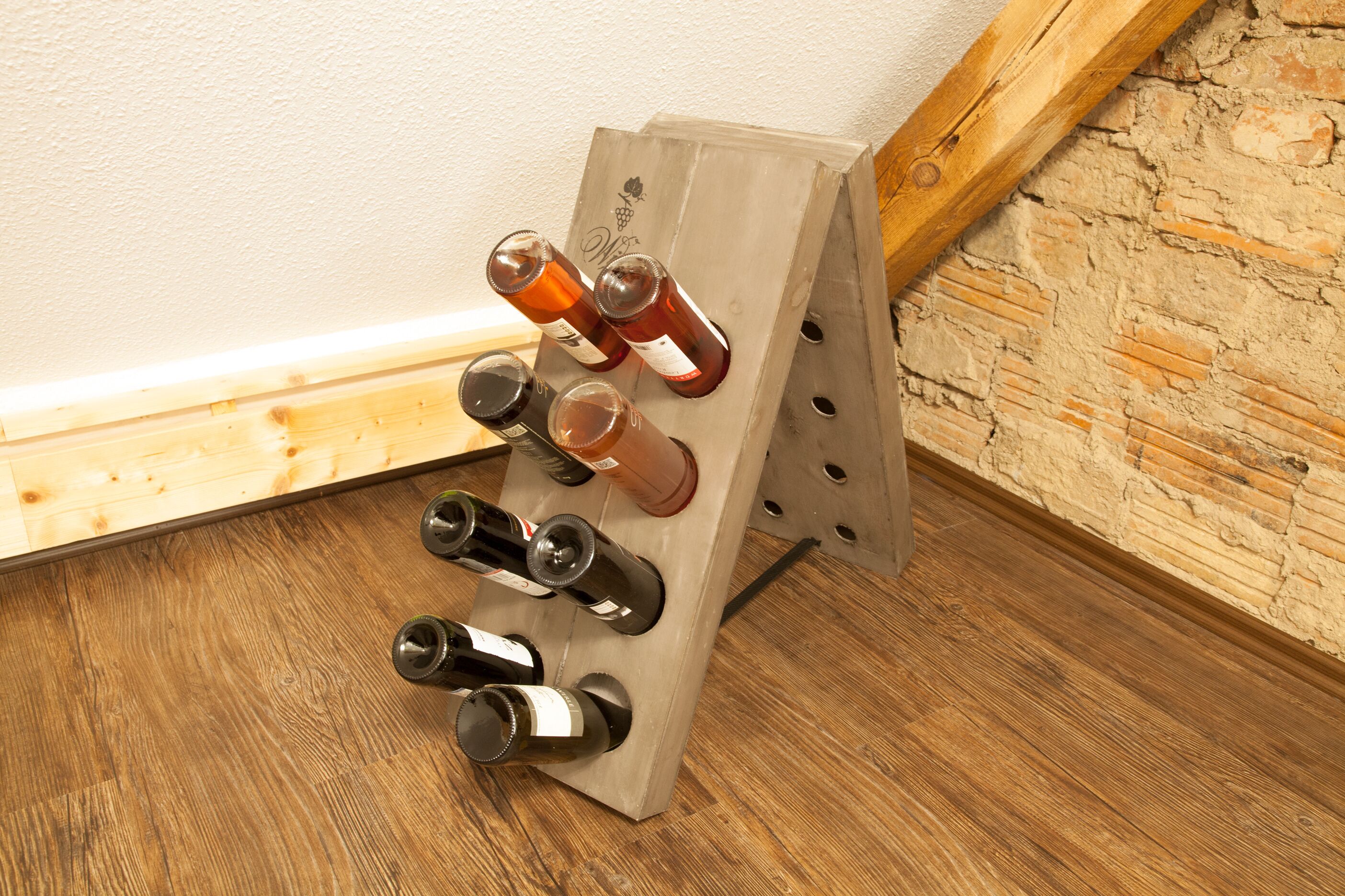 Un meuble à bouteilles, avec des bouteilles de vin, est placé dans un coin d’une pièce dont le sol est en bois et un des murs est en briques.