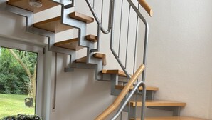 Una escalera moderna con escalones de madera y barandillas de metal, que conduce a una plataforma con un ventanal con vistas a un jardín.