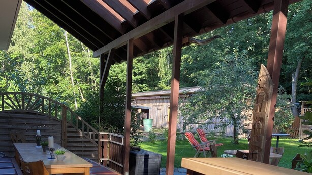 Un patio al aire libre con mobiliario de madera, incluyendo una mesa y sillas, bajo una pérgola.