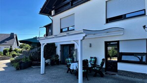 Un patio couvert avec des sièges d’extérieur, une pergola et une vue sur une maison à l’extérieur blanche et aux volets colorés.