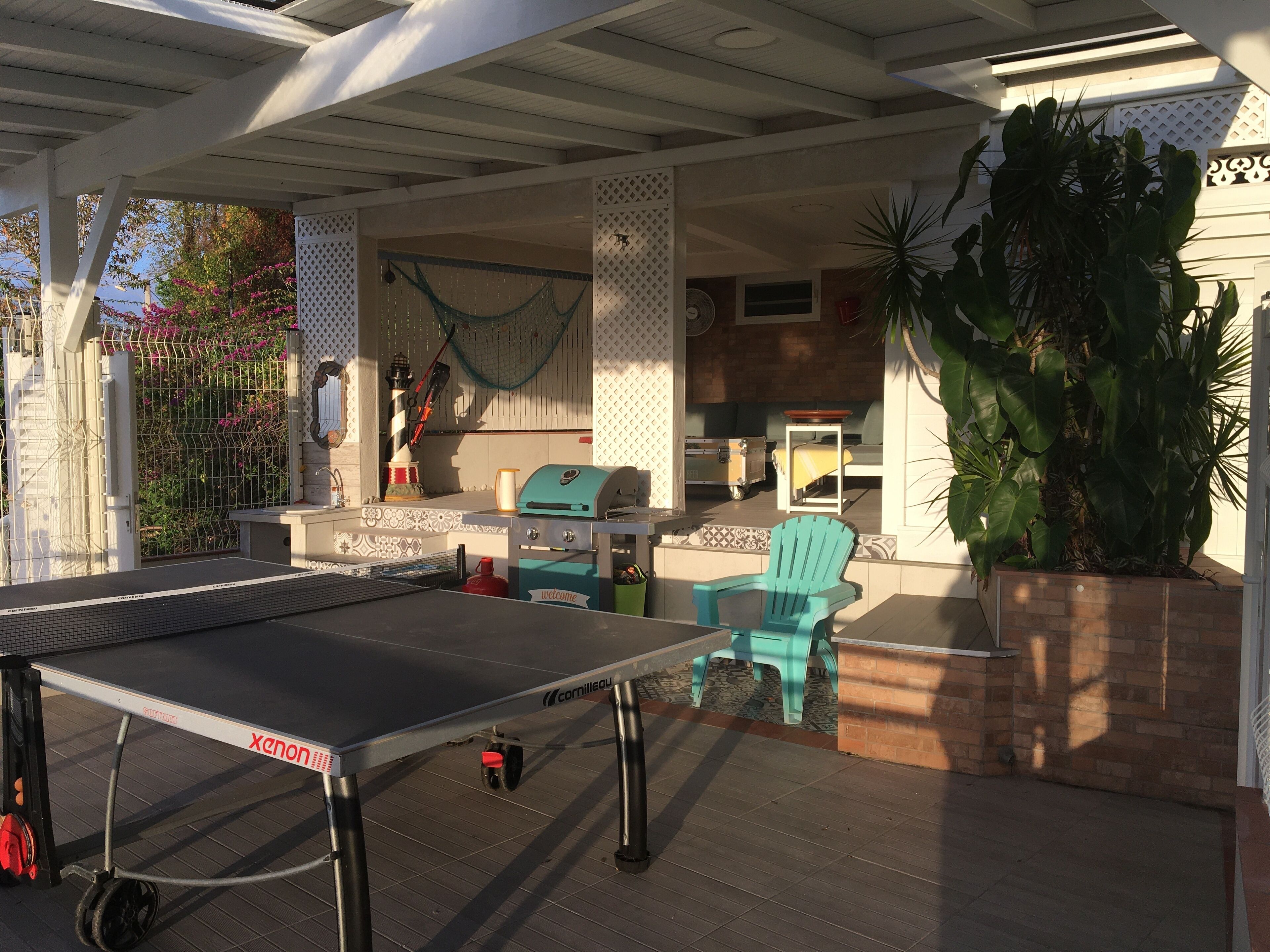 Un espace extérieur abrité comprenant une table de ping-pong, un barbecue et un coin salon avec une chaise et une petite table.
