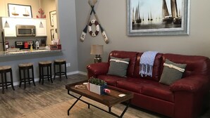 A living room with a red sofa, a coffee table, and a kitchen area with a microwave and stools. -  Village by the Beach!Townhome Walking distance to the BEACH, Heated Resort Pool (Corpus Christi)