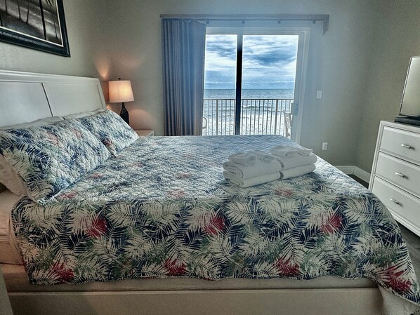 A bedroom with a bed, a nightstand, a lamp, and a view of the beach.