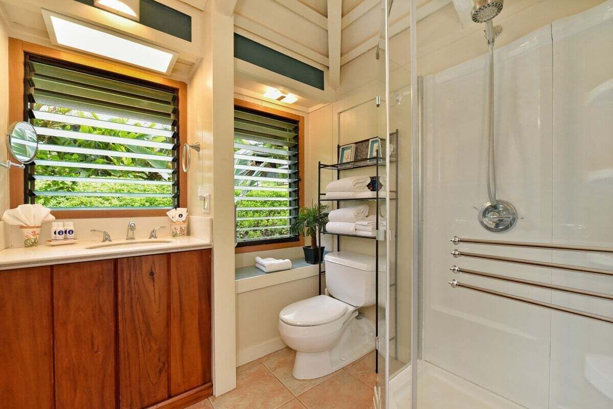 Une salle de bain avec un meuble-lavabo en bois, des toilettes, une douche et une fenêtre avec des stores.
