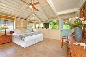 Ein Schlafzimmer mit Bett, Deckenventilator, Nachttisch, Kommode, Fenster mit Jalousien und einer Tür mit Blick auf Grünfläche.