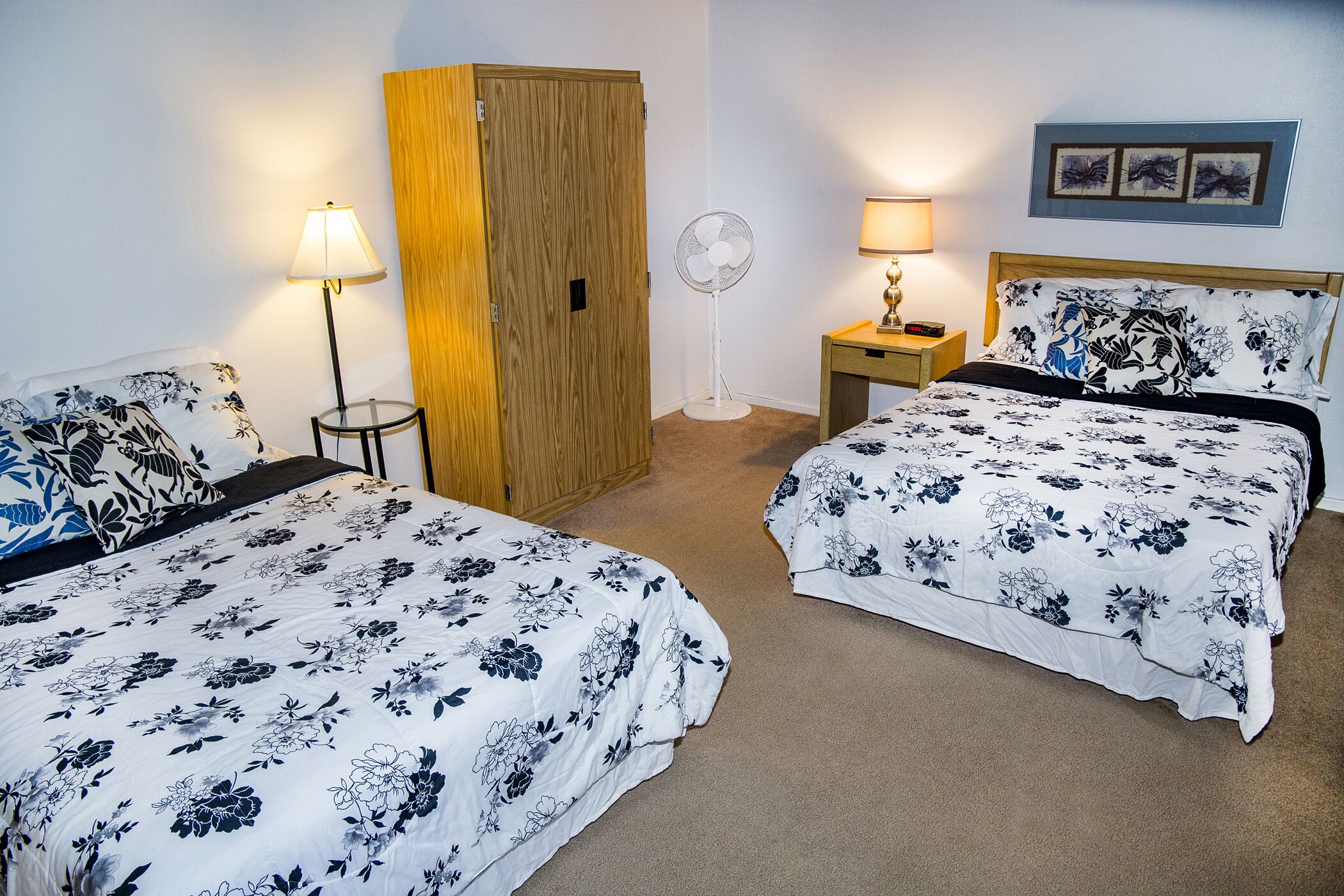 Habitación de hotel con dos camas, un armario de madera, un ventilador de pie, una mesita de noche con una lámpara y un cuadro colgado en la pared.