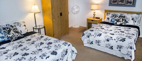 Habitación de hotel con dos camas, un armario de madera, un ventilador de pie, una mesita de noche con una lámpara y un cuadro colgado en la pared.