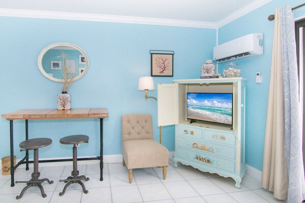 A room with a blue wall, a wooden table with two stools, a beige armchair, a television displaying a beach scene, a white air conditioner, and a decorative cabinet with drawers. - Steps to Beach Access. Boutique Bungalow Chic. (Juno Beach)