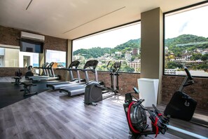 Une salle de sport équipée de tapis de course, de vélos d’appartement et d’un rameur, avec une vue sur la ville grâce à de grandes fenêtres.