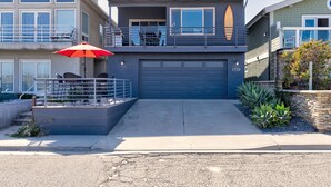 A modern two-story house with a garage, a balcony with a surfboard, and a red umbrella.