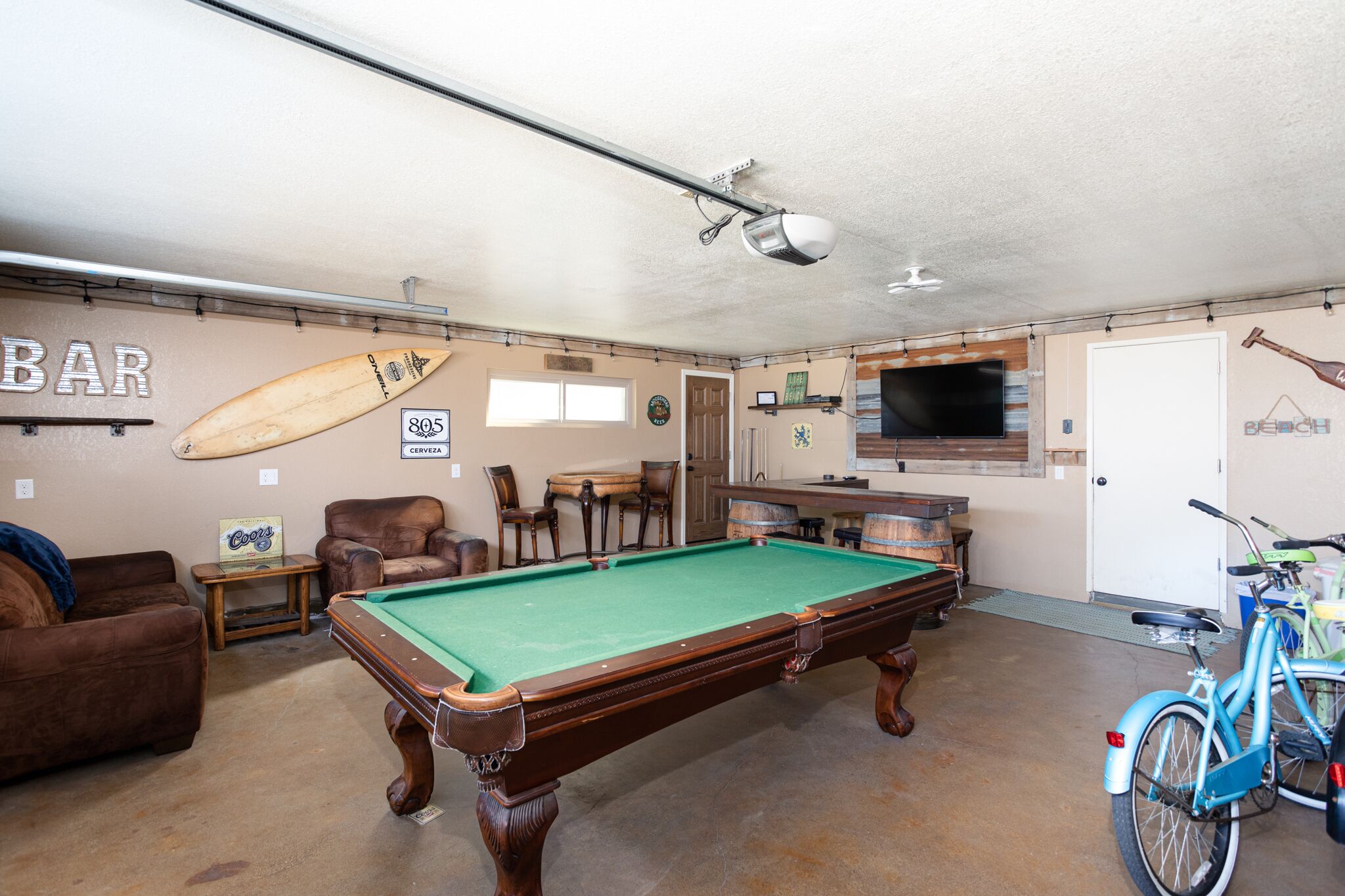 A pool table in a room with a bar area, a surfboard, and a bicycle.