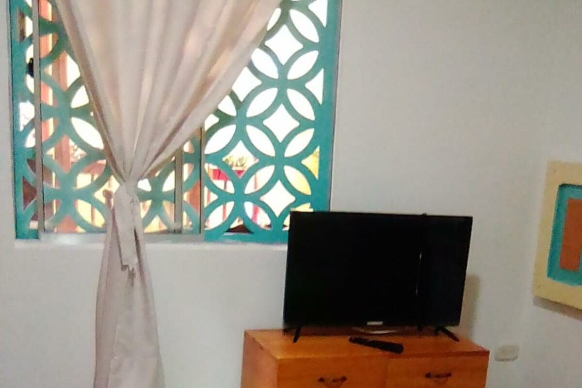 A room with a bed, a wooden TV stand, a window with a curtain, and a green bucket.