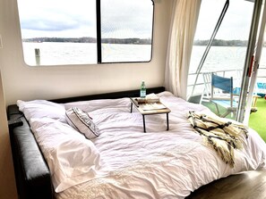 A bed with white linens, a small table with a bottle and a book, and a view of a body of water through a window.