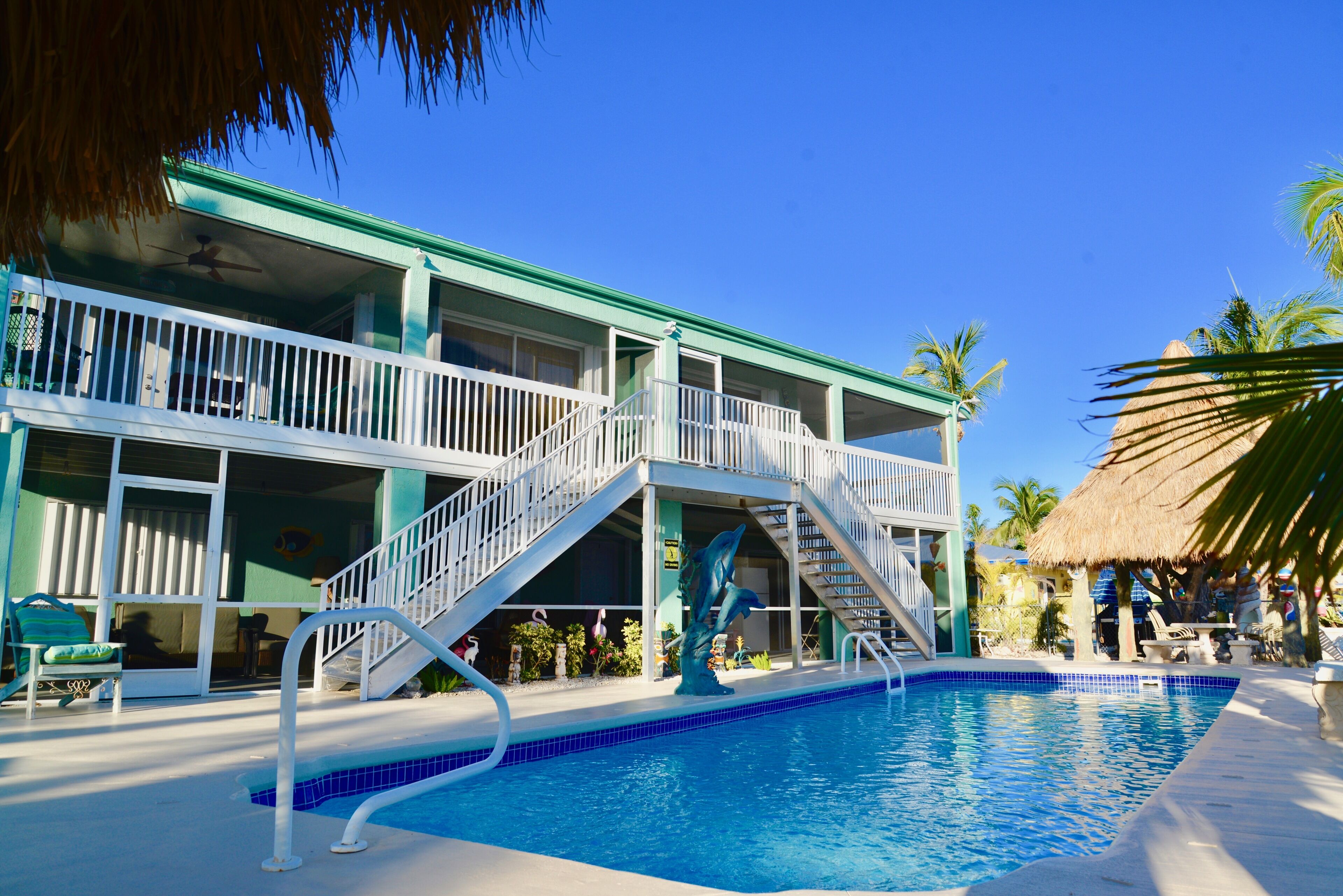 A two-story hotel with a swimming pool, outdoor seating, and a thatched-roof structure.