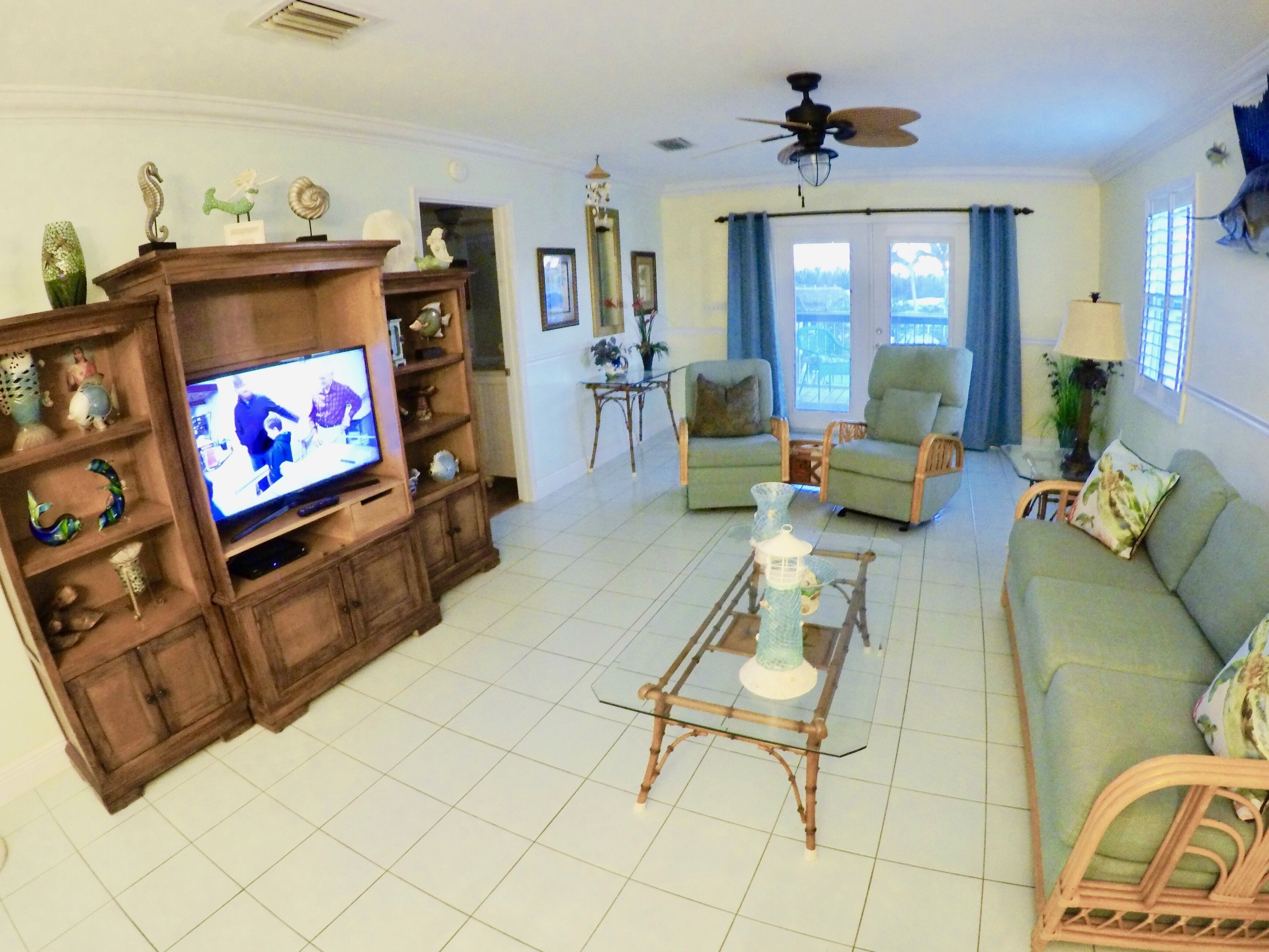 A living room with a television, two green armchairs, a glass coffee table, and a ceiling fan.