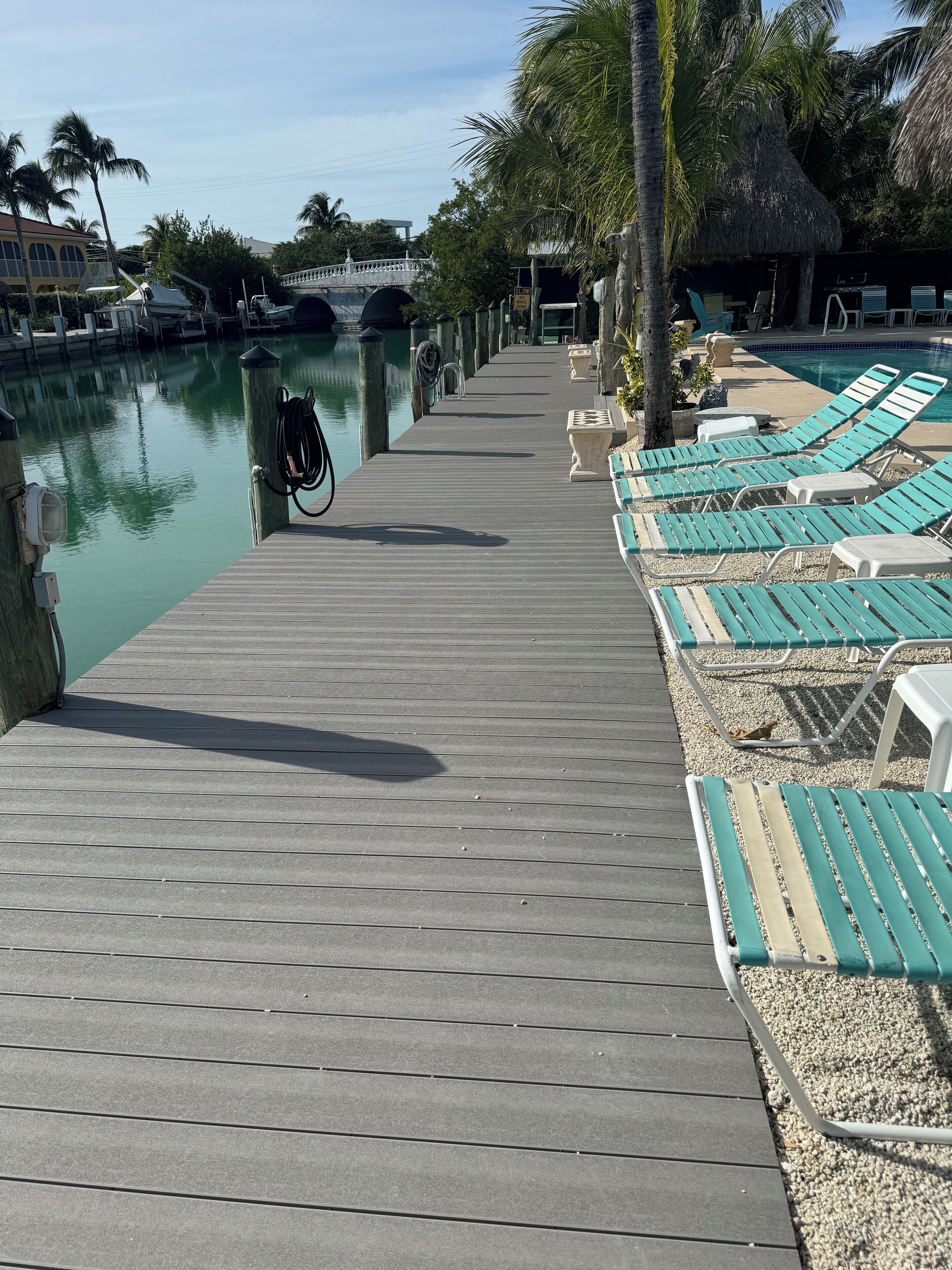 A wooden dock leading to a pool area with lounge chairs and a thatched-roof structure.