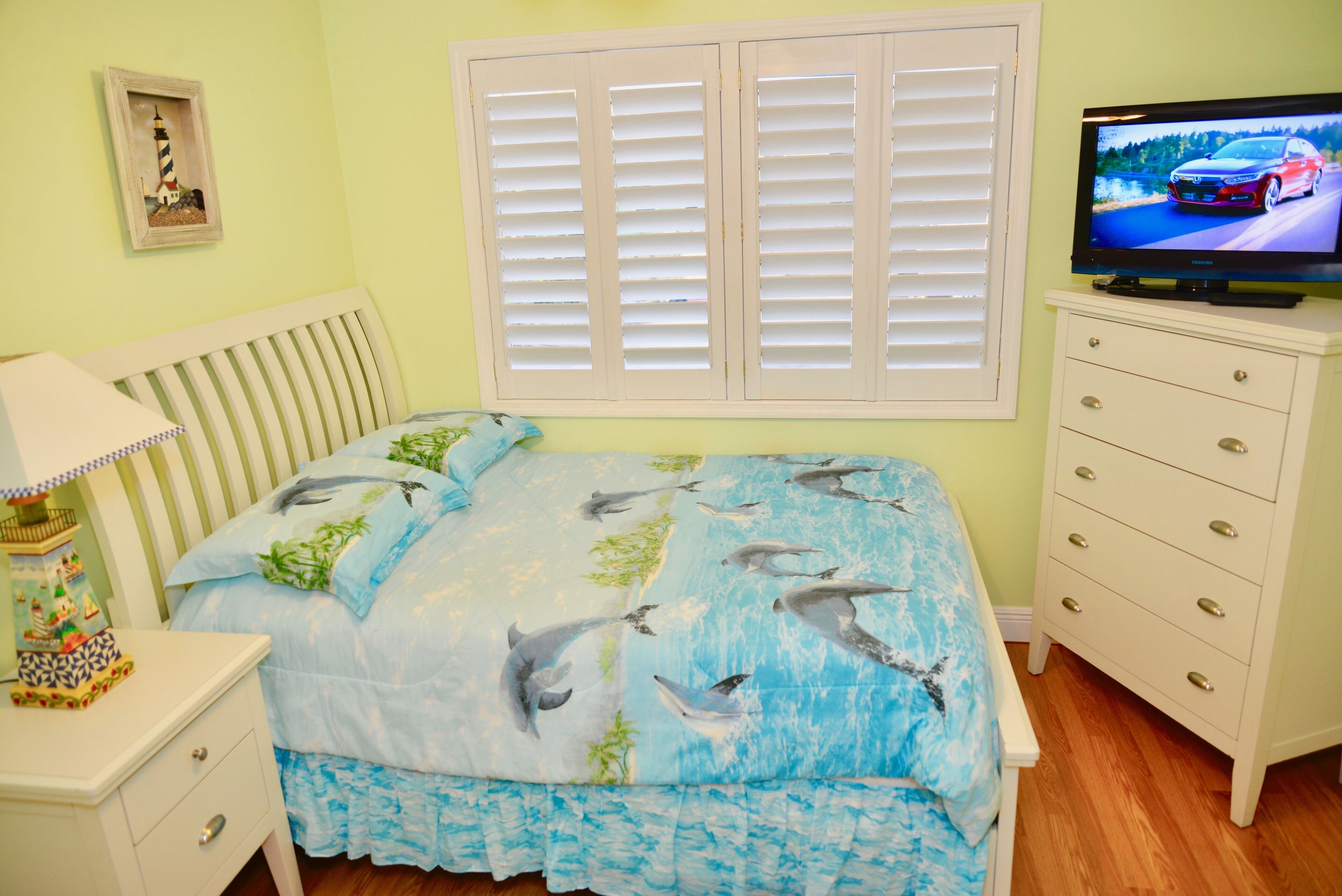 A bedroom with a bed featuring a dolphin print bedspread, a nightstand with a lamp, a dresser with a television on top, and a window with shutters.