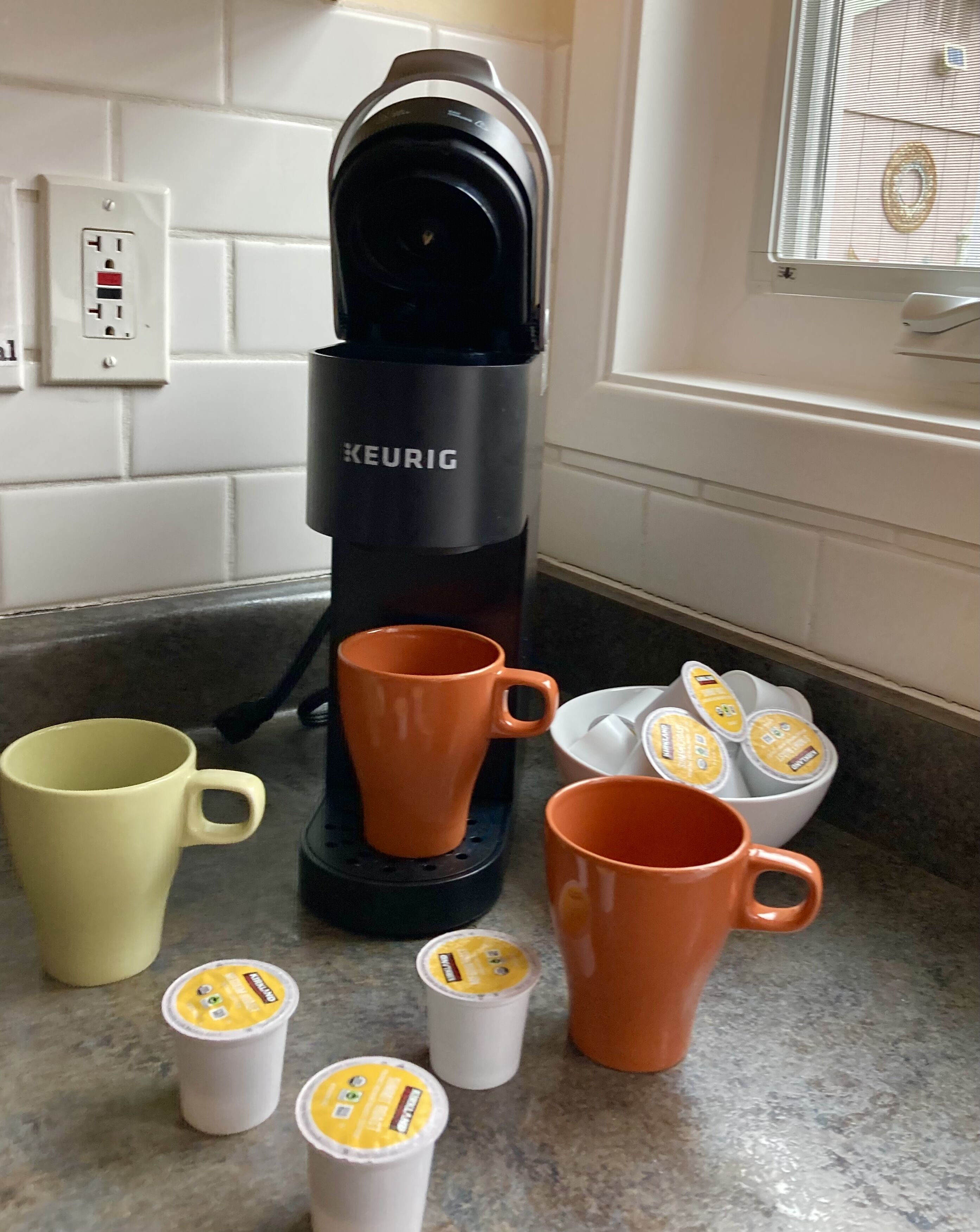 Une cuisine équipée d’une machine à café Keurig, de deux tasses oranges, d’une tasse jaune et de plusieurs dosettes de café posées sur le comptoir.