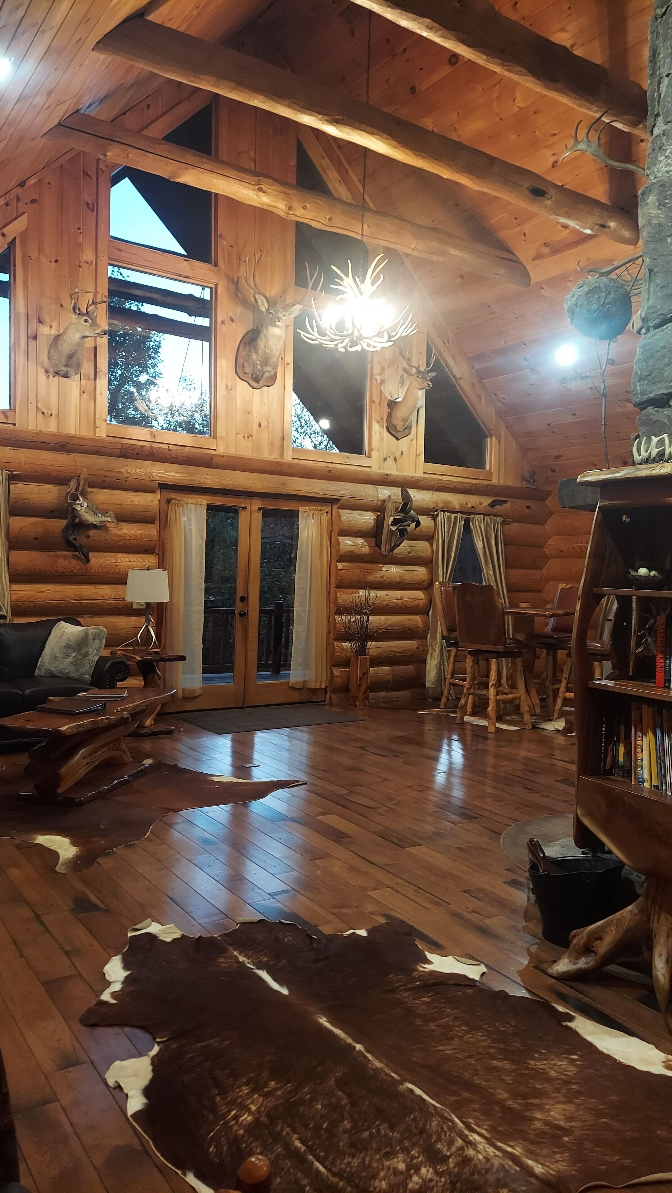 A log cabin interior with wooden walls, a chandelier, a leather rug, and mounted animal heads.