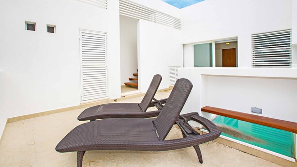 Een moderne, overdekte lounge met twee donkere wicker lounge fauteuils, een houten bank en een zwembad met glazen omheining.