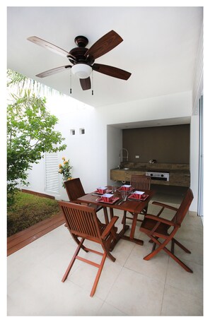 An outdoor dining area with a table and chairs, a ceiling fan, and a kitchen area in the background.