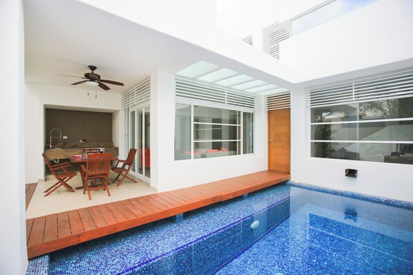 A modern outdoor pool area with a wooden deck, a dining table with chairs, and a ceiling fan.
