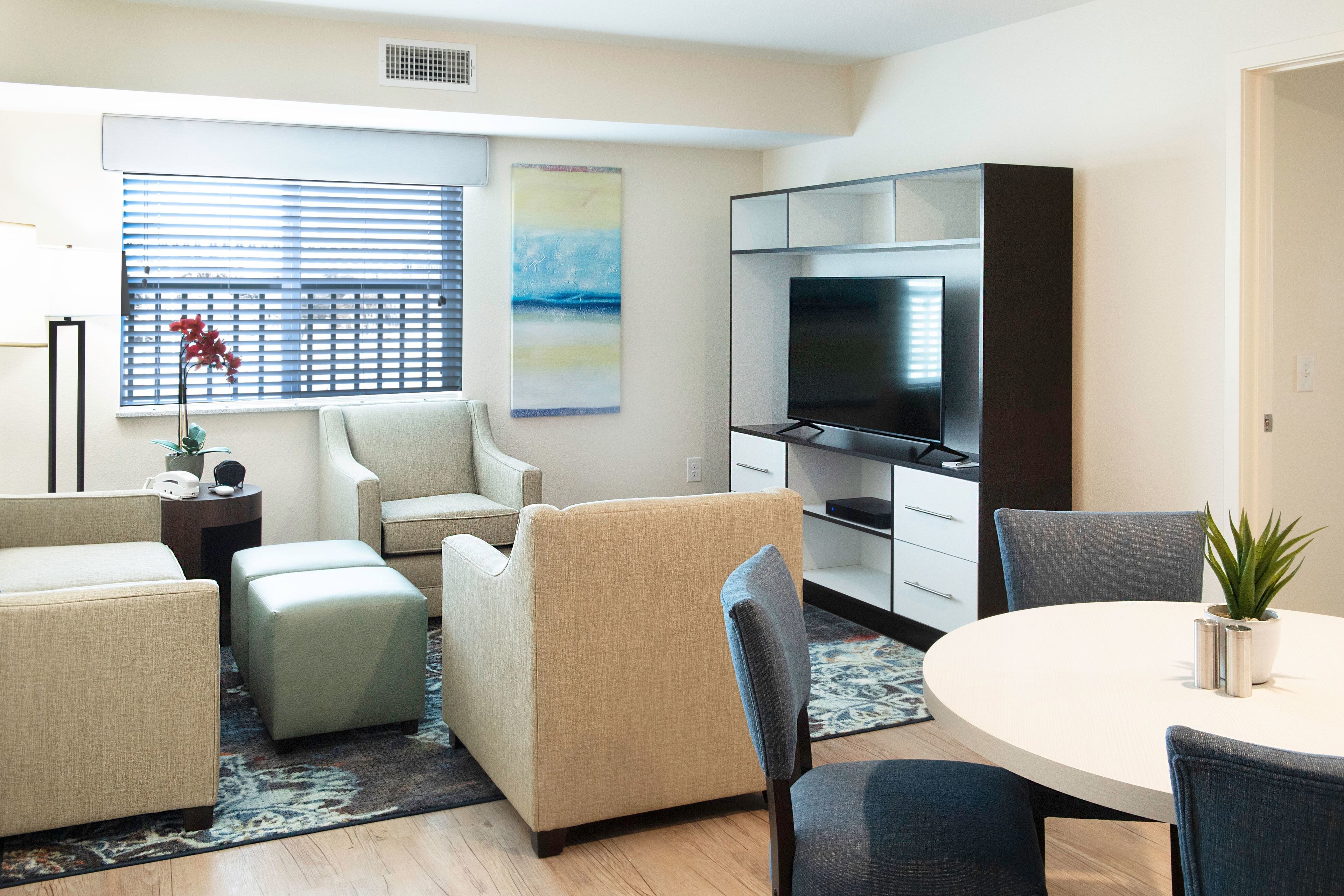 A modern living room with a flat-screen TV, beige sofas, a dining table with chairs, and a large window with blinds.