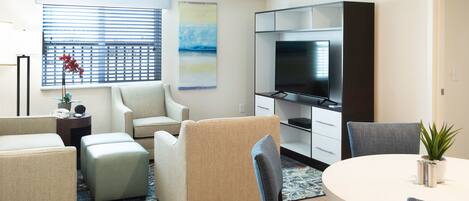 A modern living room with a flat-screen TV, beige sofas, a dining table with chairs, and a large window with blinds.
