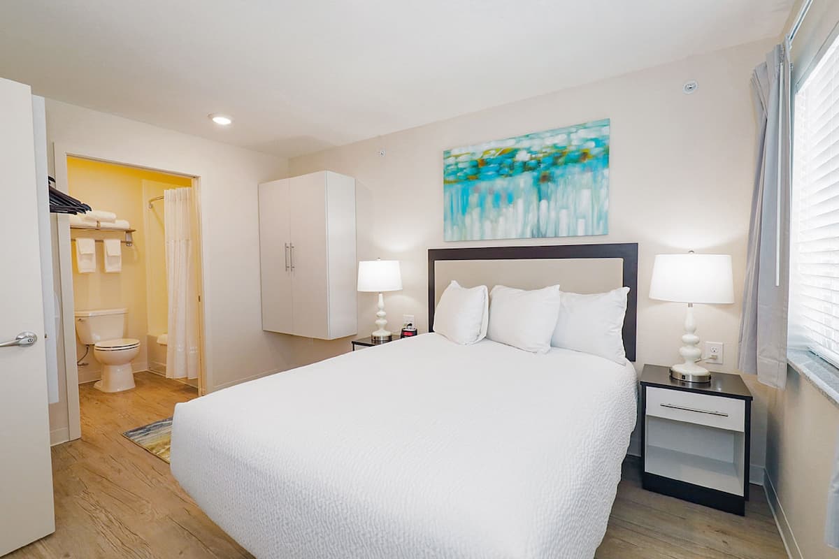 A hotel room with a large bed, two bedside tables with lamps, a painting on the wall, and a bathroom visible through an open door.
