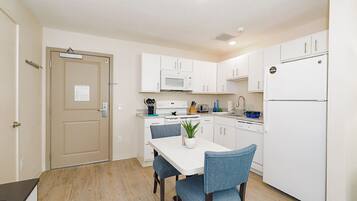 A compact kitchen with white cabinets, a microwave, a stove, and a refrigerator. There is a dining table with two chairs and a potted plant.