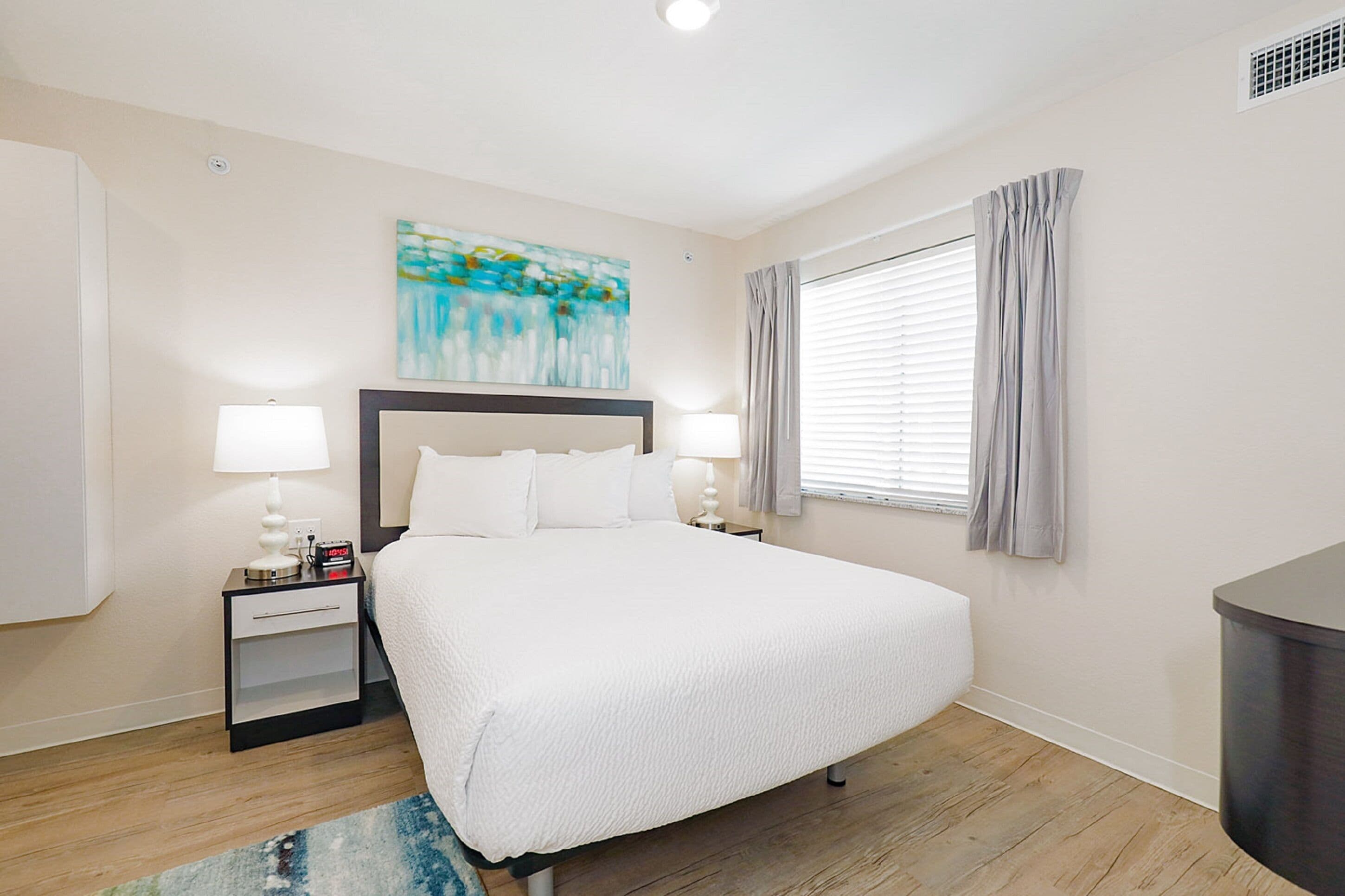 A hotel room with a large bed, two bedside tables with lamps, a painting on the wall, and a window with curtains.