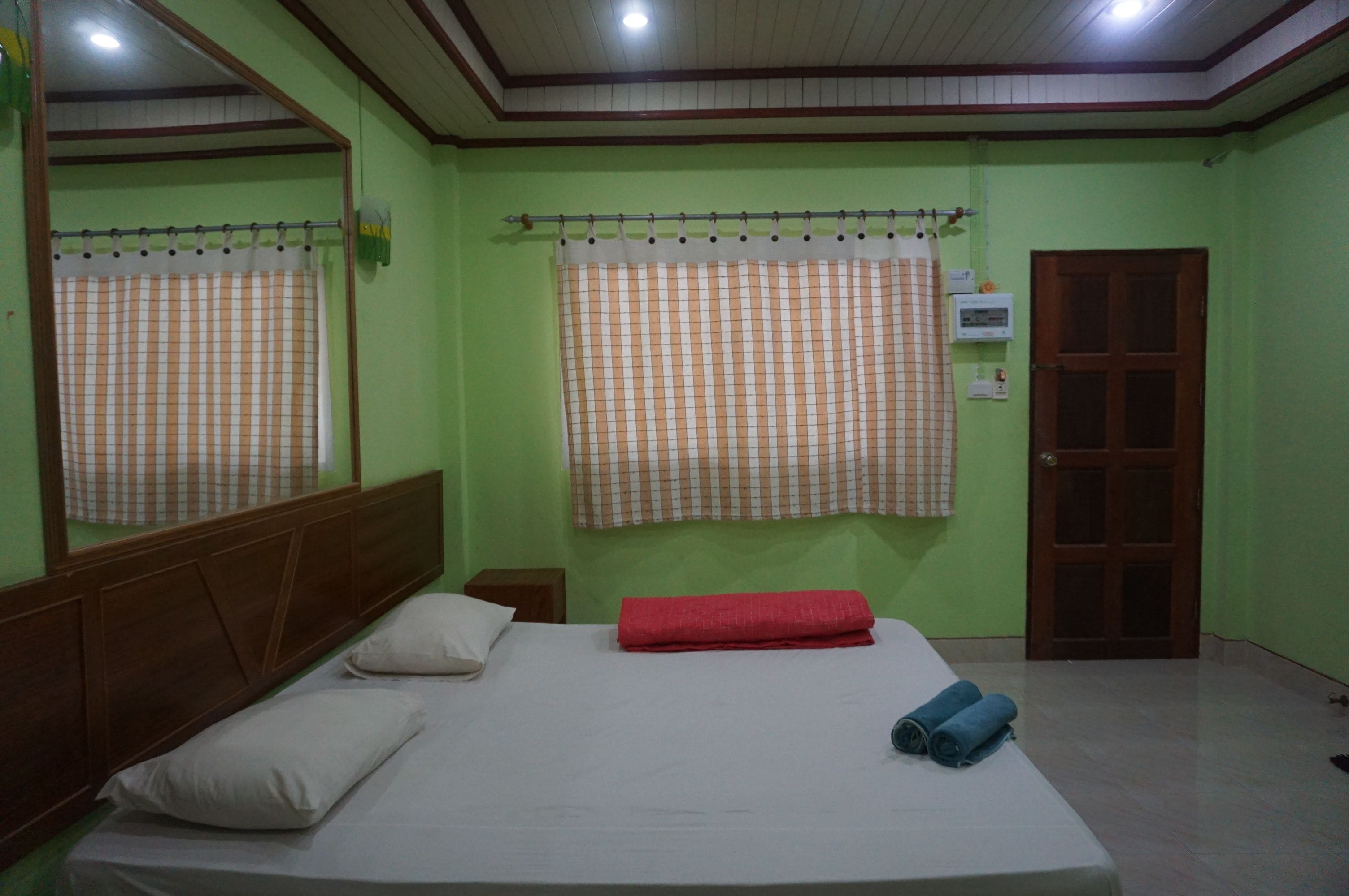 Quarto com uma cama de solteiro, cabeceira de madeira, toalha vermelha e janela com cortina xadrez.