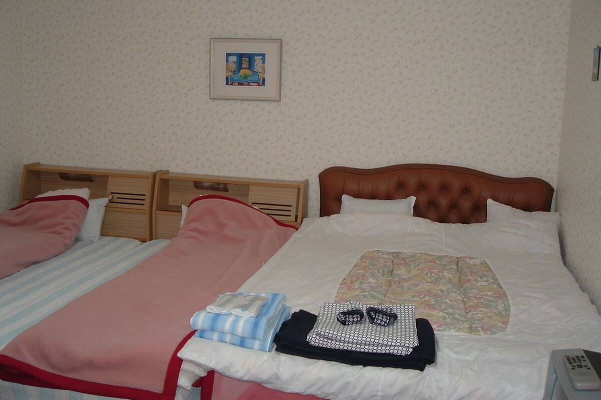 A room with two beds, one with a striped bedspread and the other with a floral bedspread. There is a framed picture on the wall, a bedside table with a lamp, and a small table with a remote control.