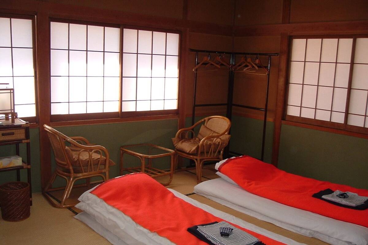A traditional Japanese room with tatami flooring, two low beds with red and white bedding, a wooden chair, a small table, and a hanging clothes rack.