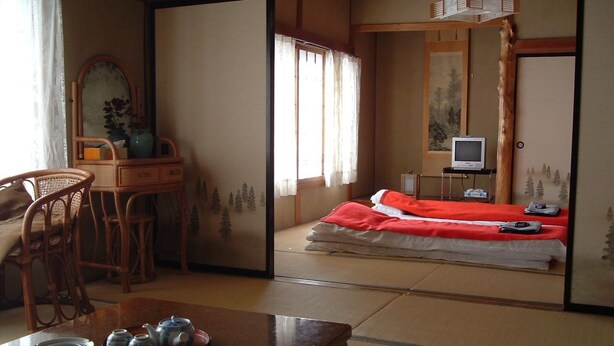 Une pièce de style japonais traditionnel, avec un sol recouvert de tatamis, une petite table avec un service à thé, un lit avec une literie rouge et un téléviseur posé sur un meuble.