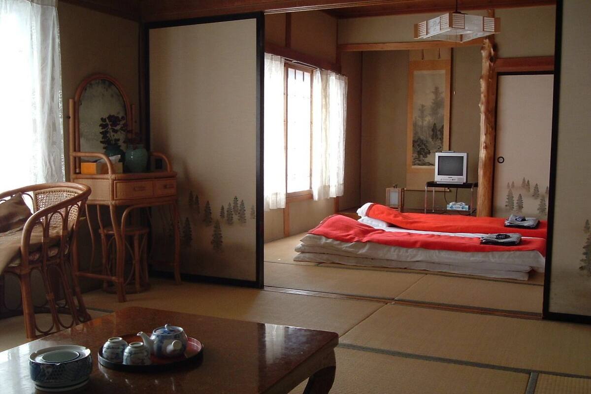 A traditional Japanese room with tatami flooring, a low table with tea set, a bed with red bedding, and a television on a stand.