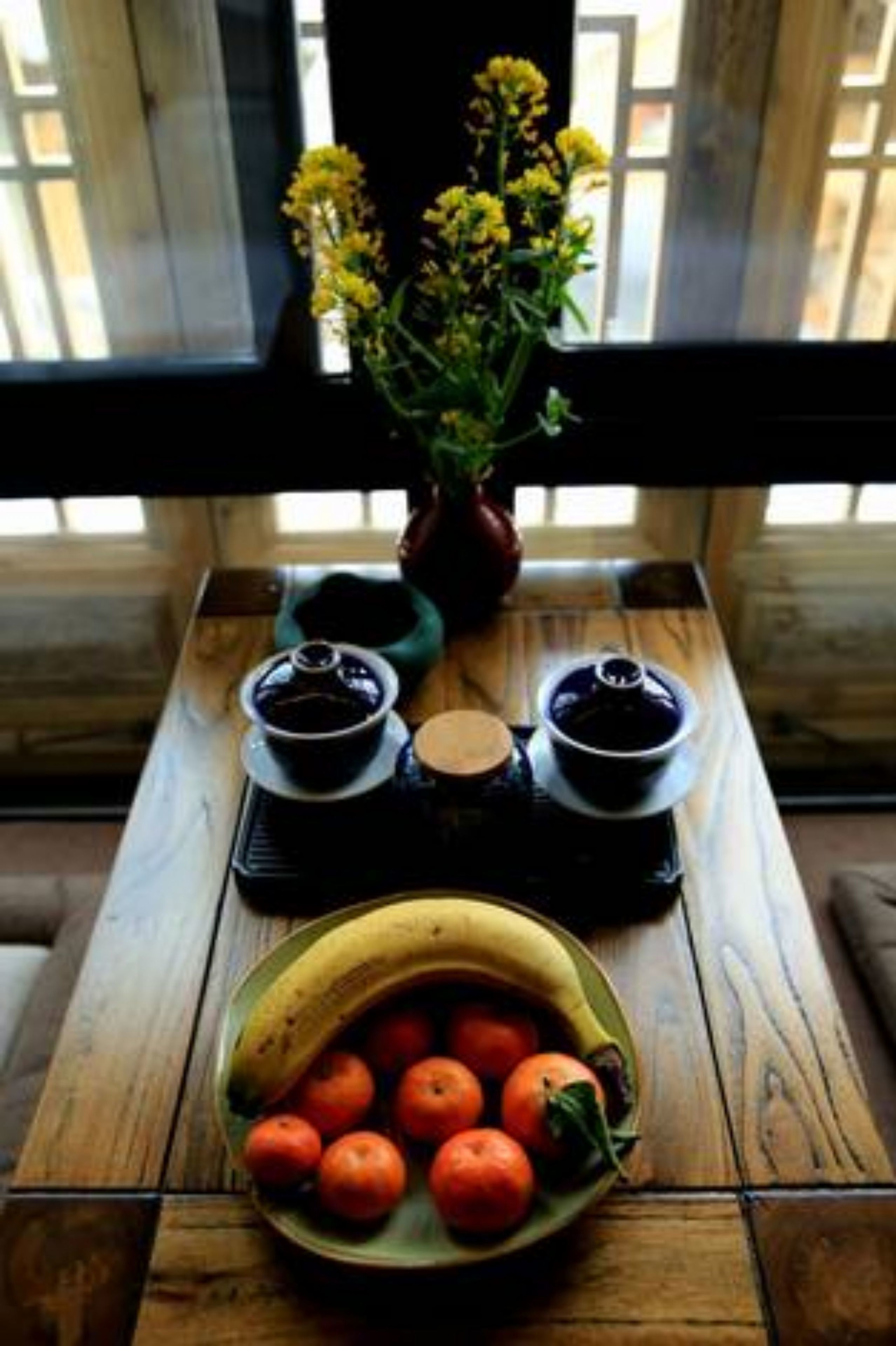 A wooden table with a bowl of oranges and a vase of yellow flowers.