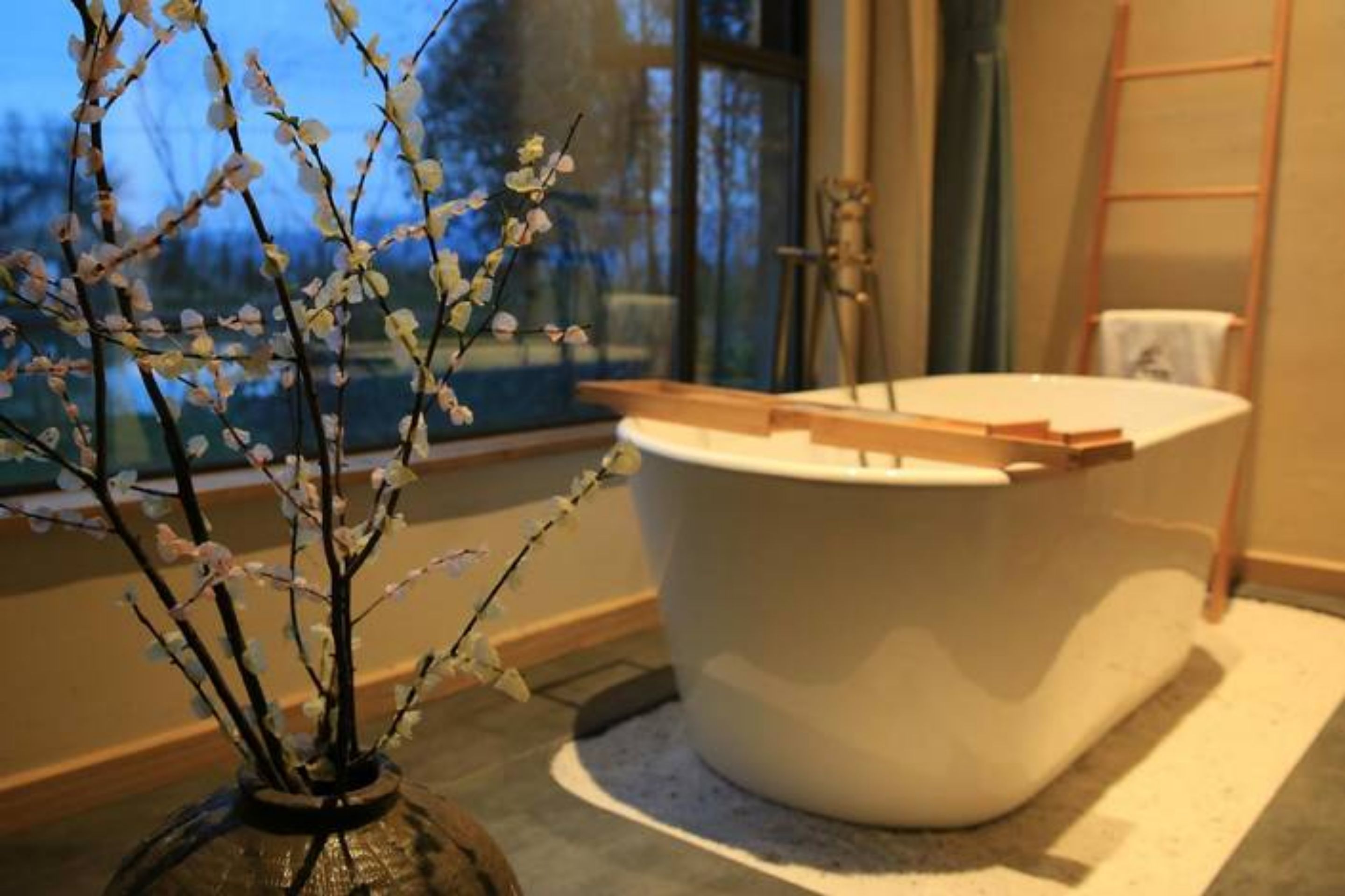 A modern bathroom with a freestanding bathtub, a window with a view of trees, and a ladder against the wall.