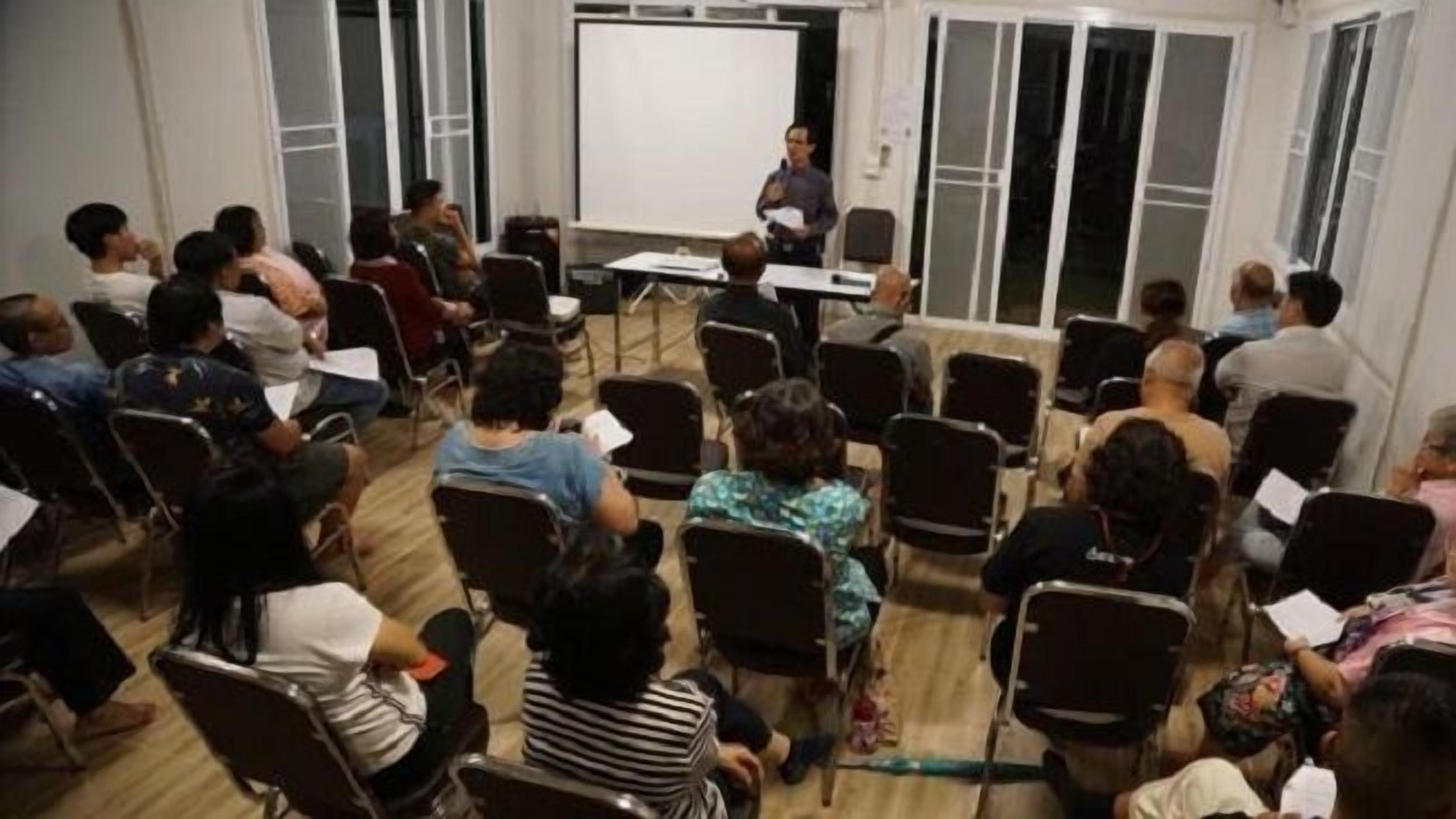 Un salón de conferencias con una pizarra, sillas dispuestas en filas, y asistentes mirando hacia el frente.