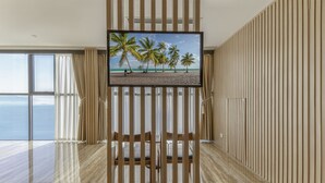 Ein modernes Zimmer mit einem großen Fernseher auf einem Holzboden, Blick auf den Strand durch große Fenster und ebenfalls ein Holzboden.