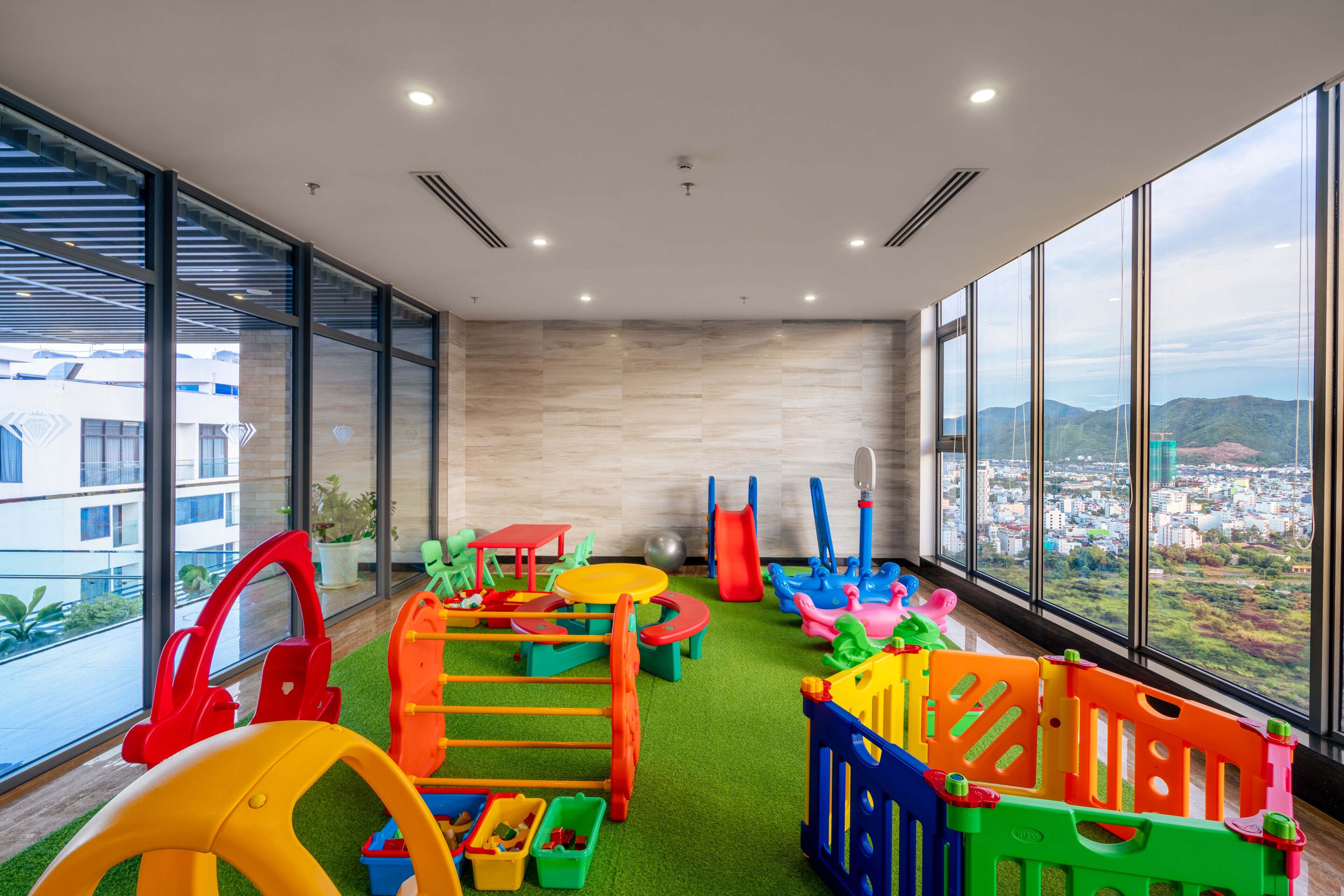Un espace de jeux pour enfants avec divers jouets et structures ludiques, de grandes fenêtres offrant une vue sur la ville et un aménagement intérieur moderne.