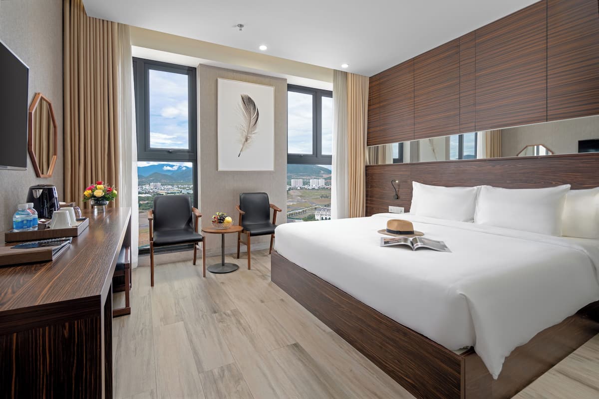 Et moderne hotelværelse med en stor seng, et skrivebord med kaffemaskine, en stol og udsigt til byens skyline gennem store vinduer.