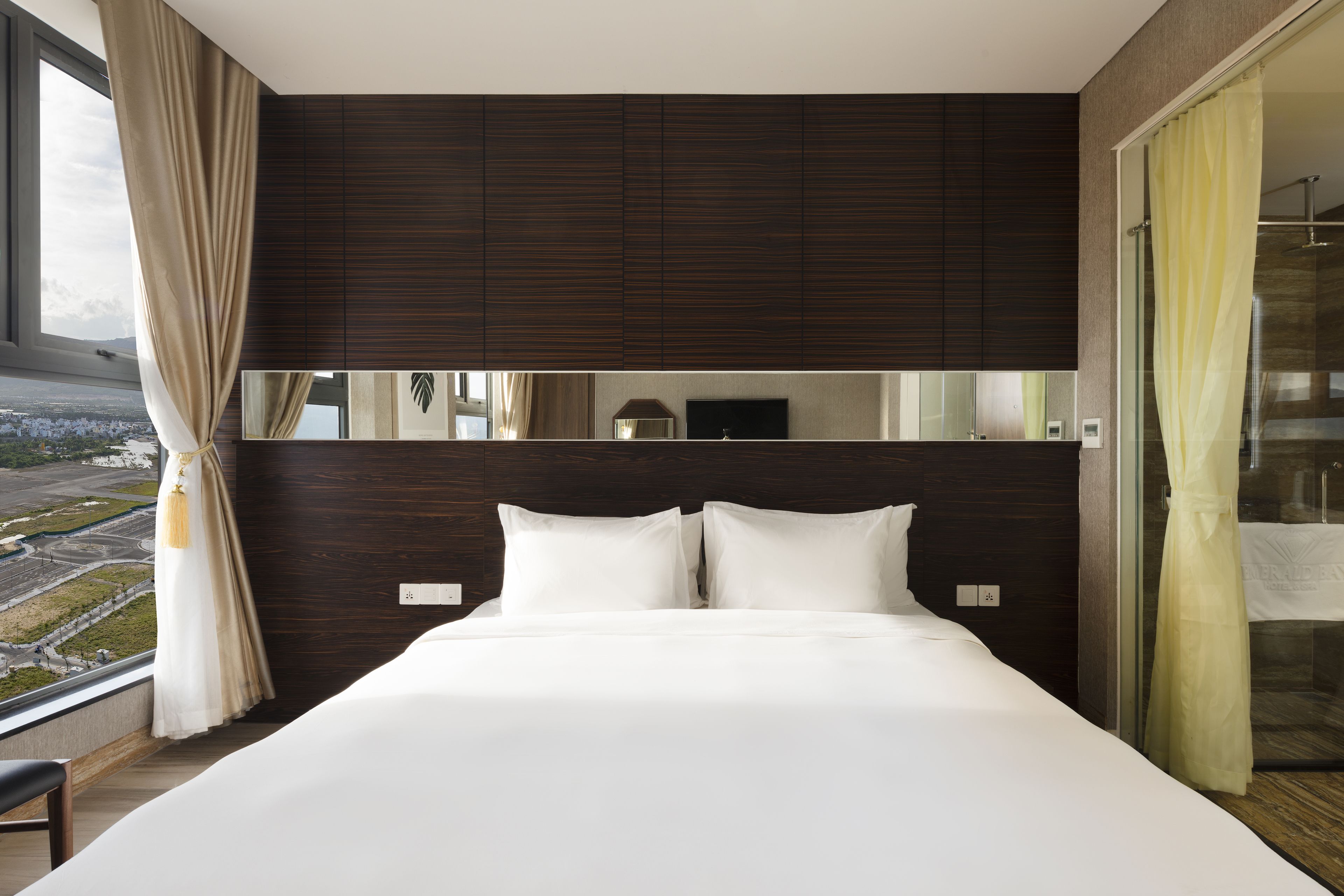 A modern hotel room with a large bed, dark wood paneling, a large window with a view, and a glass shower area.
