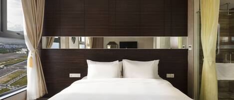 A modern hotel room with a large bed, dark wood paneling, a large window with a view, and a glass shower area.