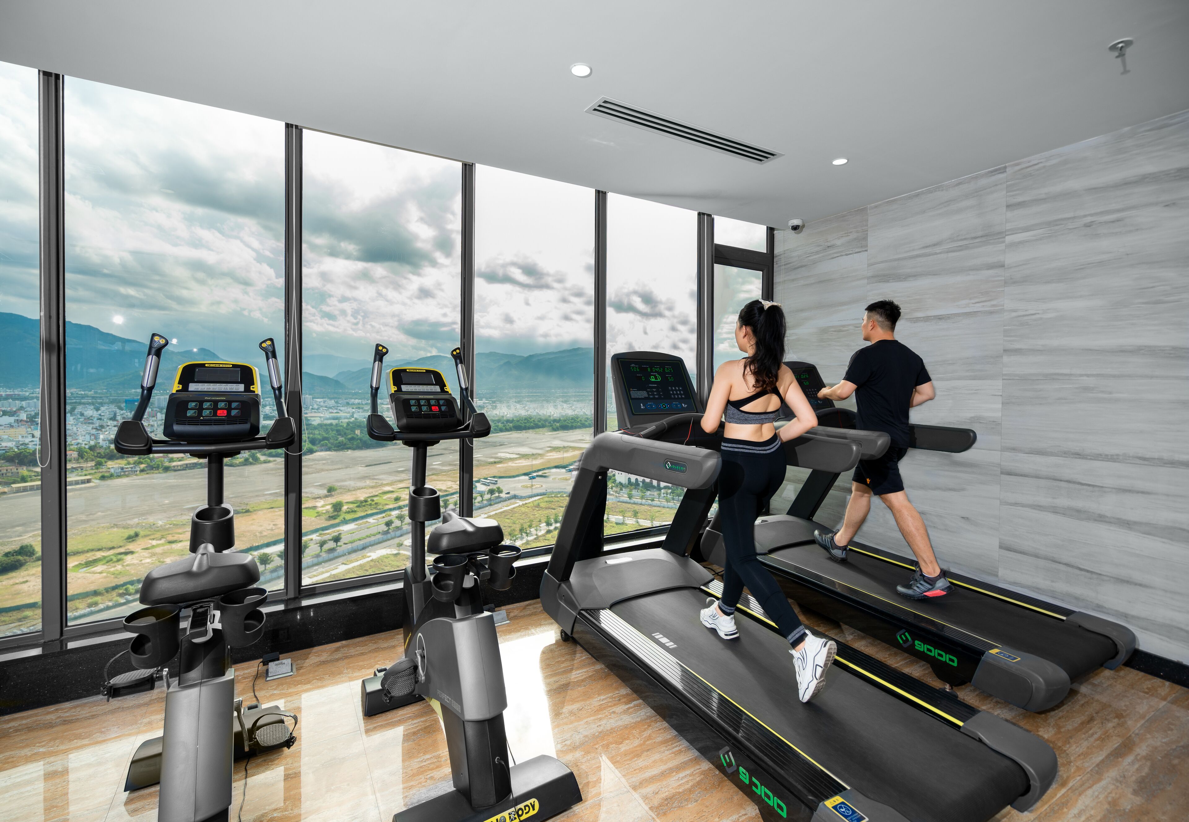 A gym with treadmills, stationary bikes, and a large window with a city view.