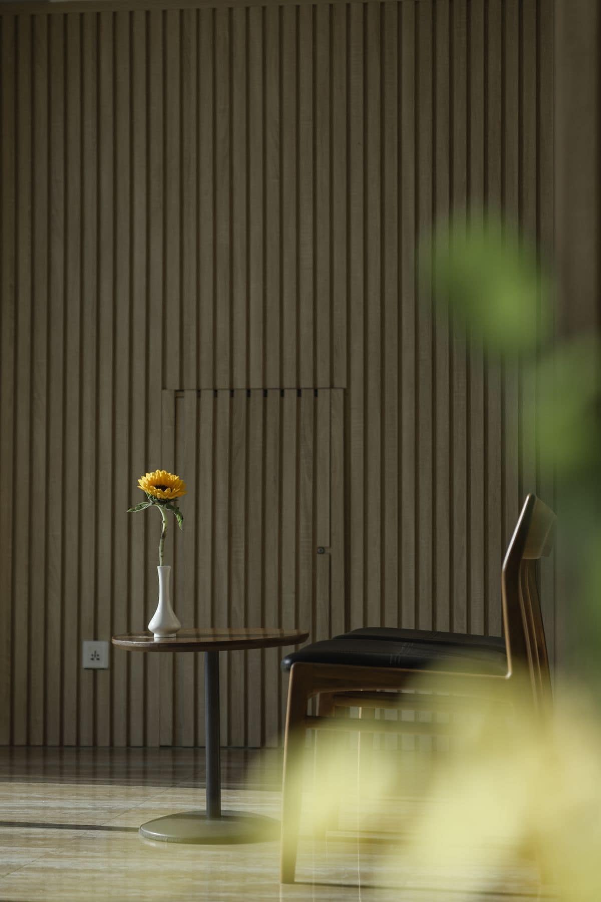 A room with a wooden slatted wall, a white vase with a sunflower, a wooden chair, and a small round table.