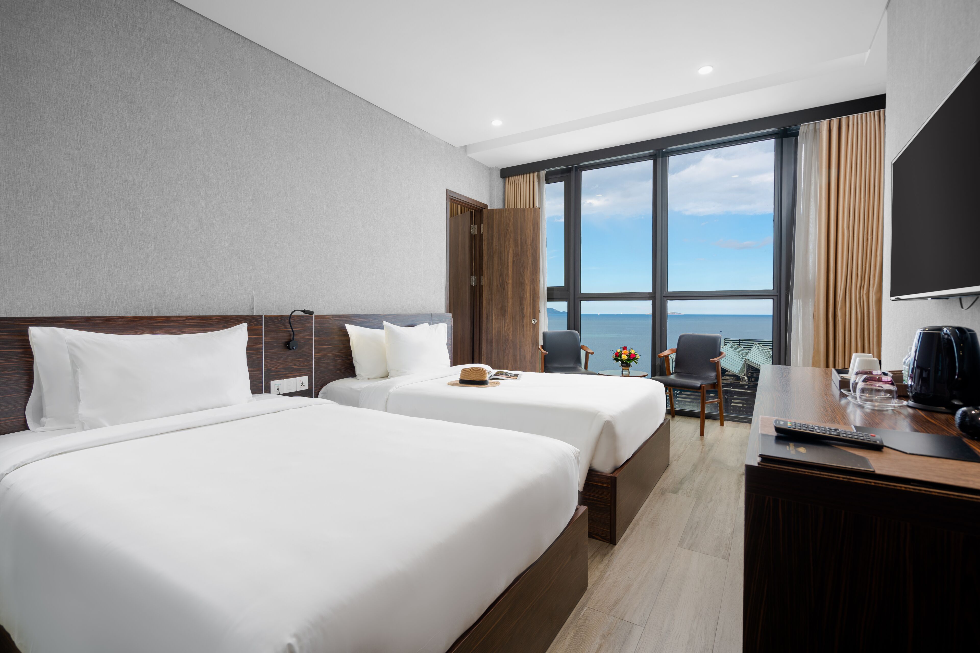 A hotel room with two beds, a desk with a coffee maker, a TV, and a view of the ocean.