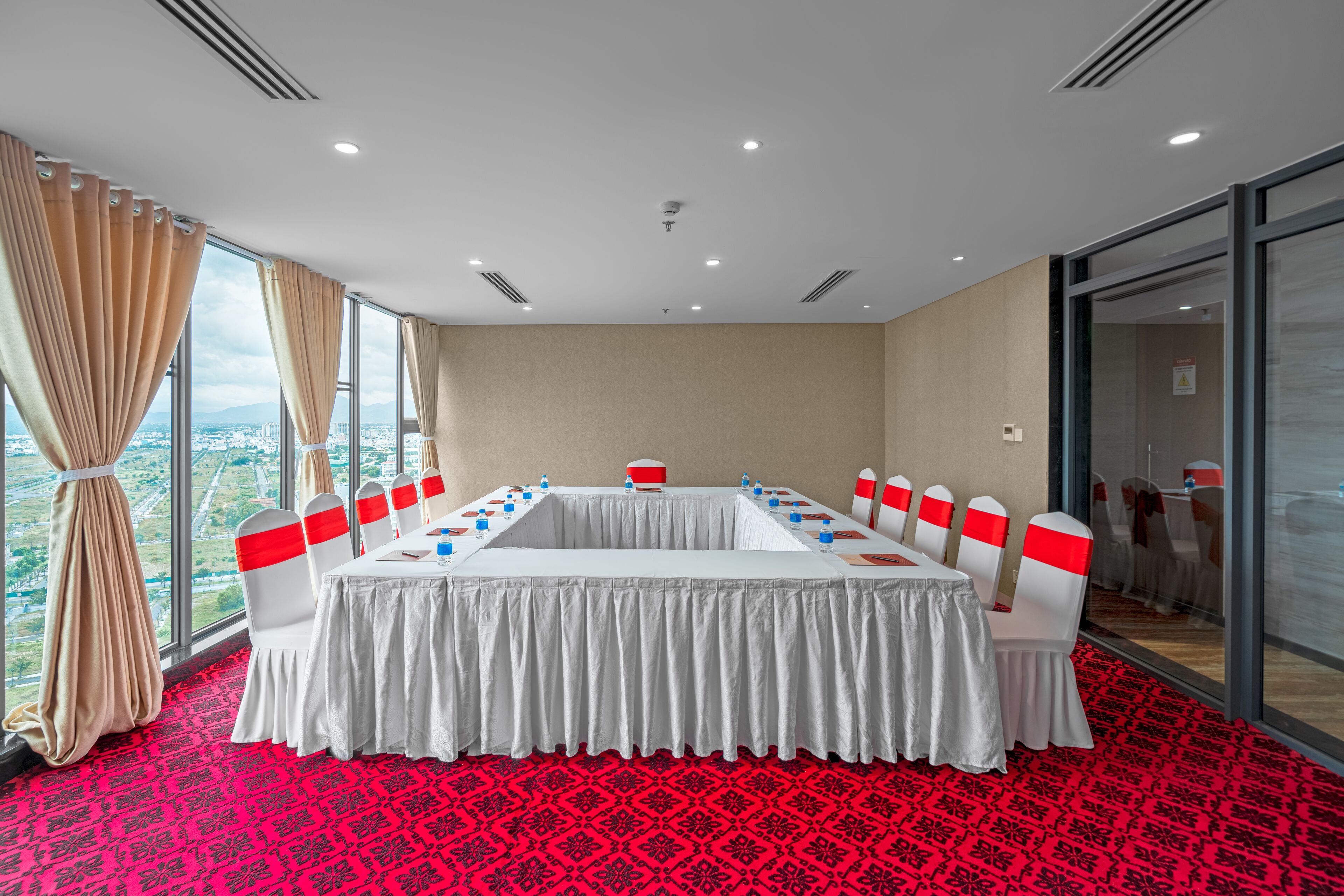 Un salón de conferencias con una larga mesa, sillas blancas con cintas rojas, una alfombra roja estampada y grandes ventanales con vista a la ciudad.