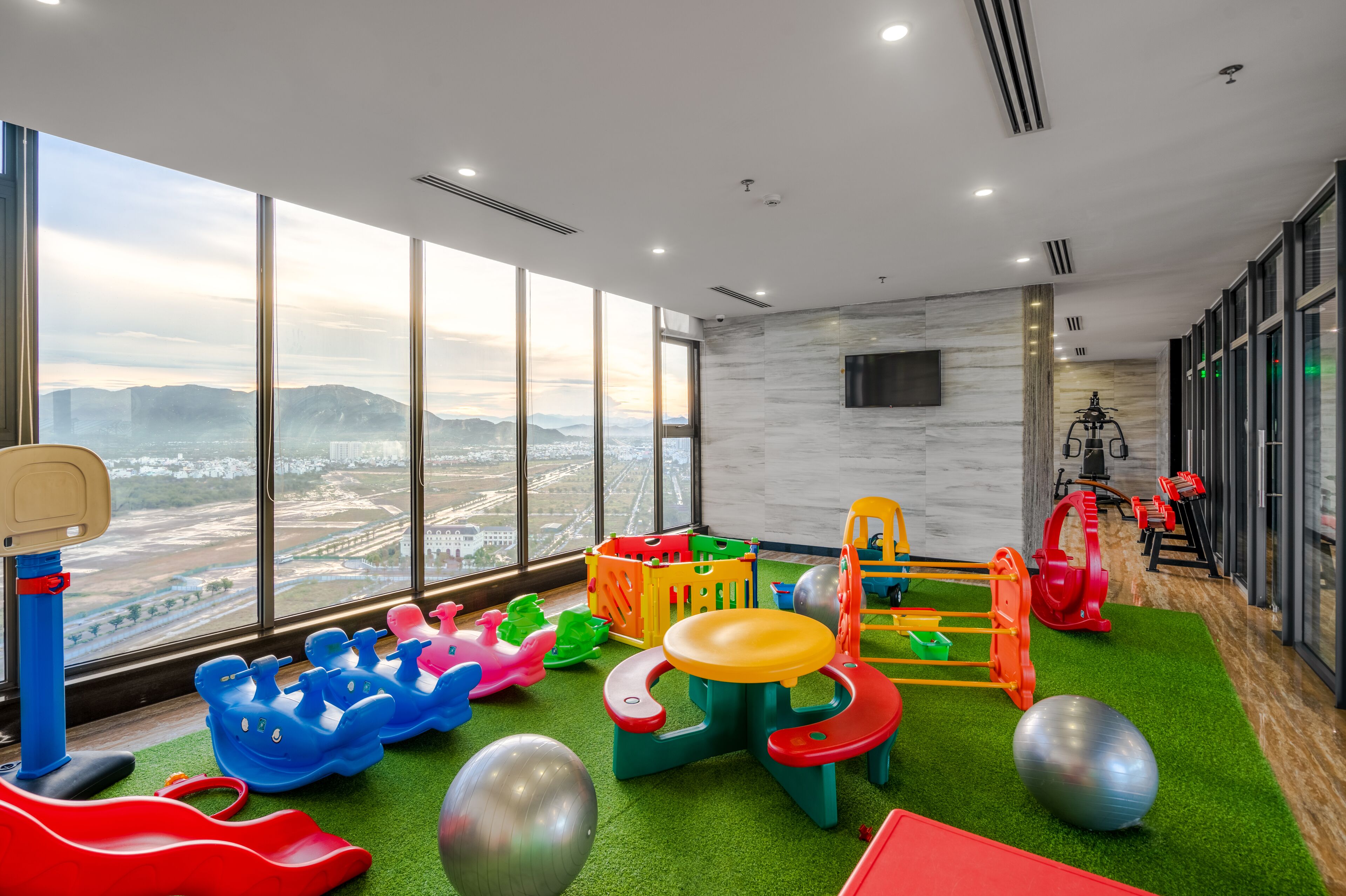 Ein Spielbereich für Kinder mit verschiedenen Spielsachen und Geräten, große Fenster mit Blick auf die Stadt und ein an der Wand befestigter Fernseher.