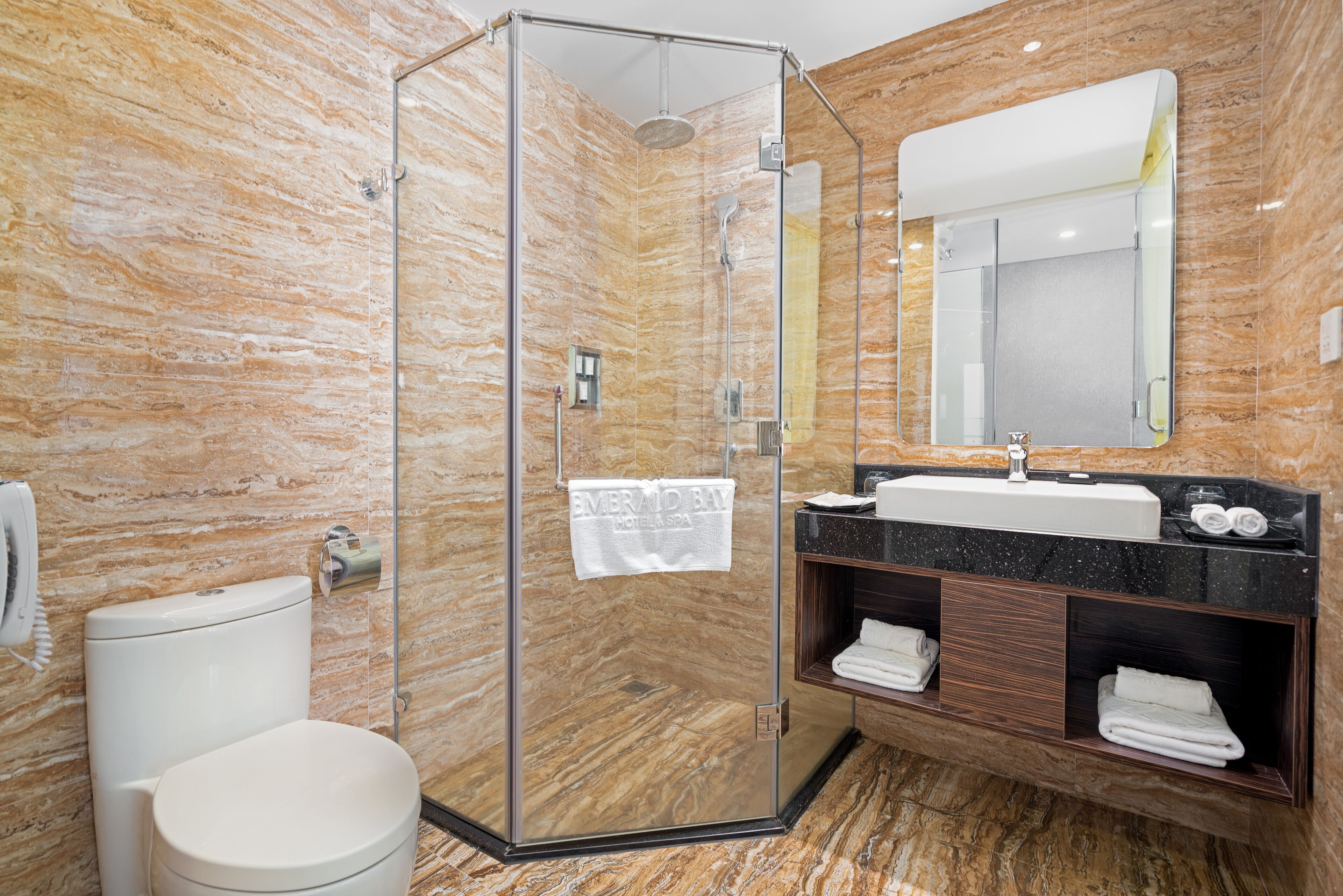 Un baño moderno con ducha envuelta en cristal, inodoro blanco, pared de mármol, lavamanos con espejo y toallas dobladas con cuidado.